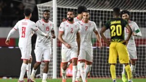 Tunisia players celebrate a goal during a match against Jamaica, featuring red and white kits, while a Jamaican player watches.
