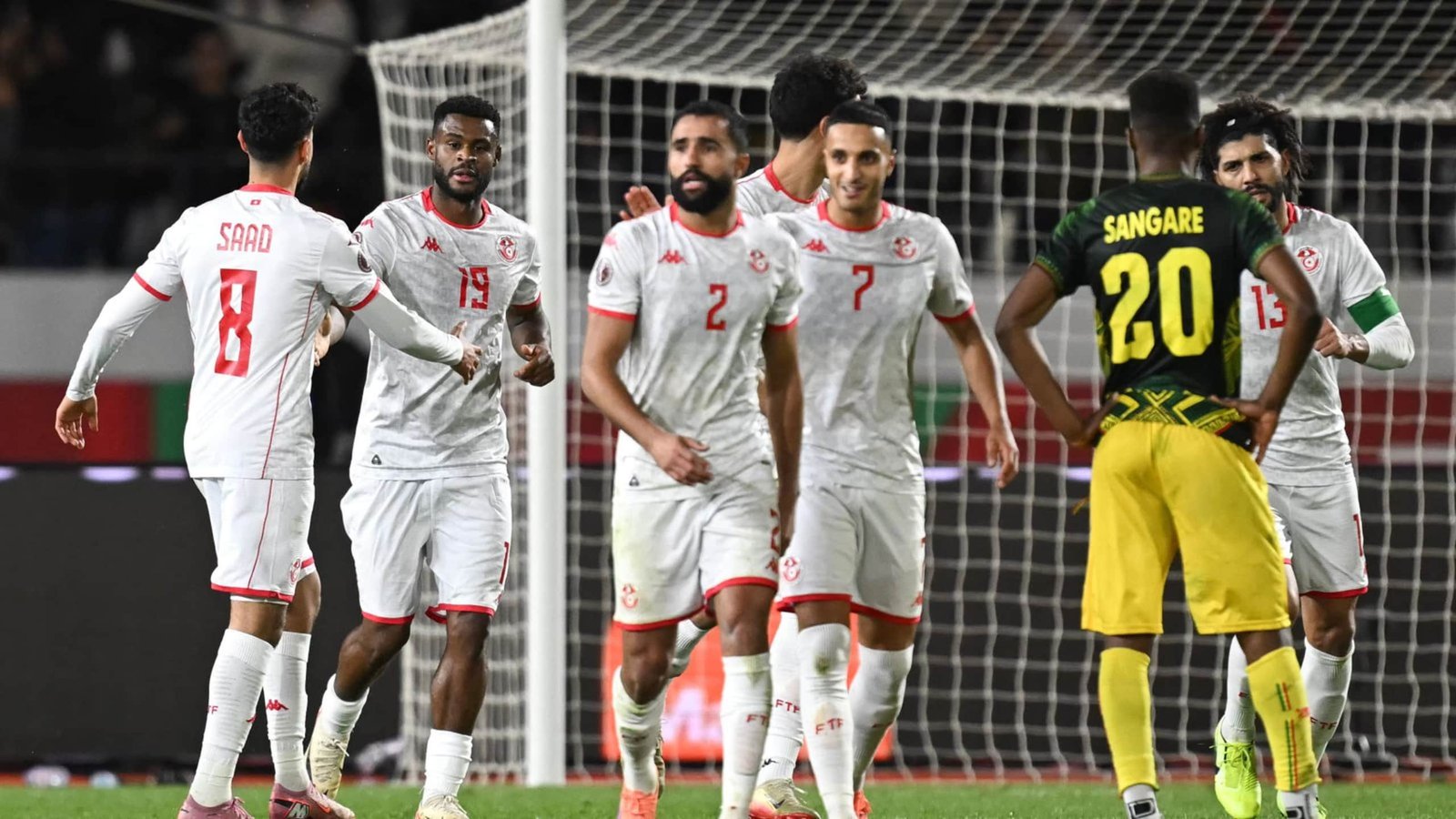 Tunisia players celebrate a goal during a match against Jamaica, featuring red and white kits, while a Jamaican player watches.