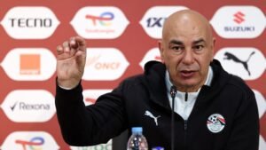 Egyptian football coach gestures while speaking at a press conference, wearing a black hoodie featuring the Egyptian FA badge, with sponsor logos in the background.