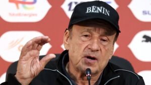 Coach addressing the media in a black cap reading "BENIN," emphasising points during a press conference backdrop featuring logos.