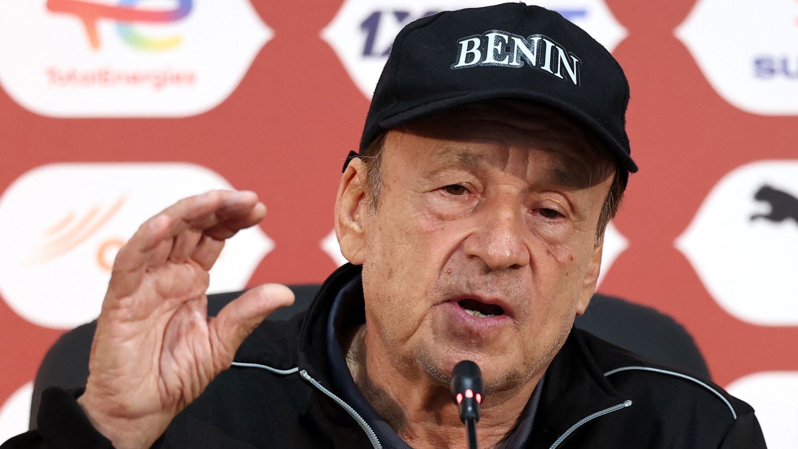 Coach addressing the media in a black cap reading "BENIN," emphasising points during a press conference backdrop featuring logos.