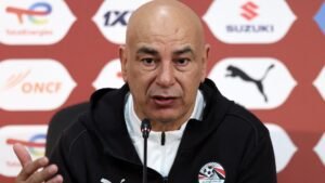 L'entraîneur de football égyptien s'exprimant lors d'une conférence de presse, portant un survêtement noir avec le logo de la Fédération égyptienne, sur fond bordeaux.