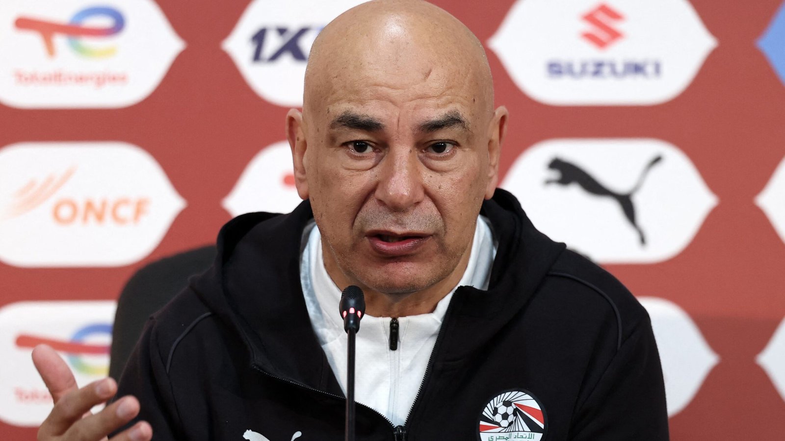 L'entraîneur de football égyptien s'exprimant lors d'une conférence de presse, portant un survêtement noir avec le logo de la Fédération égyptienne, sur fond bordeaux.