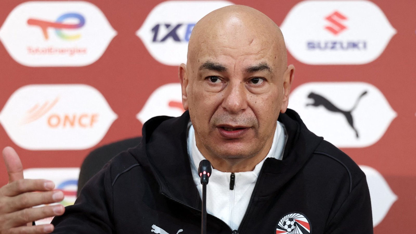 Head coach speaks at a press conference, wearing a black jacket with a club logo, amidst a backdrop featuring sponsor logos.