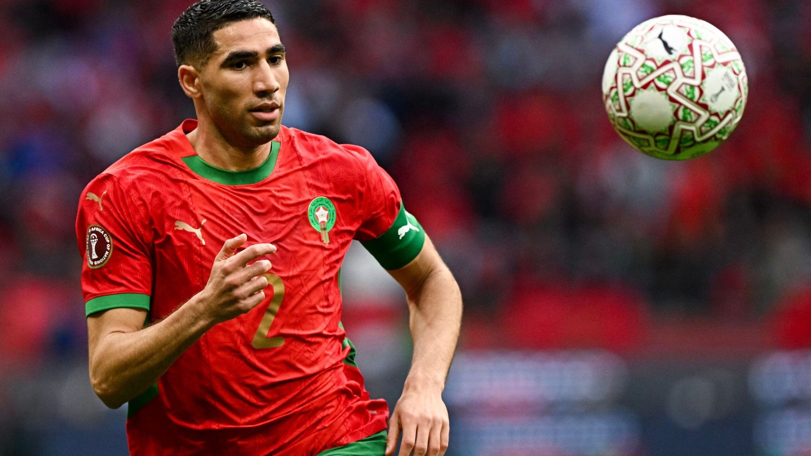 Achraf Hakimi in Morocco’s red kit, poised to receive a football during an intense match, showcasing his skills and focus.