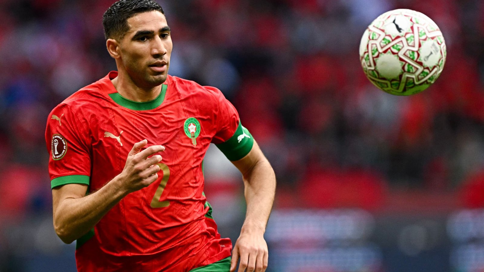 Achraf Hakimi, in Morocco's vibrant red kit with green accents, controls a football during a match at a major tournament.
