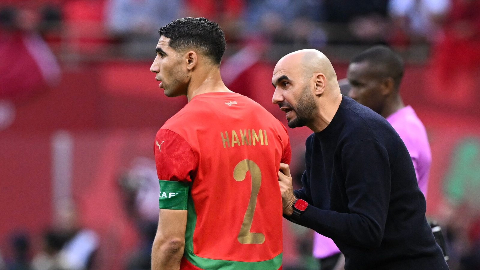 Achraf Hakimi, wearing the Moroccan national team jersey, listens intently to coach Walid Regragui during a match.