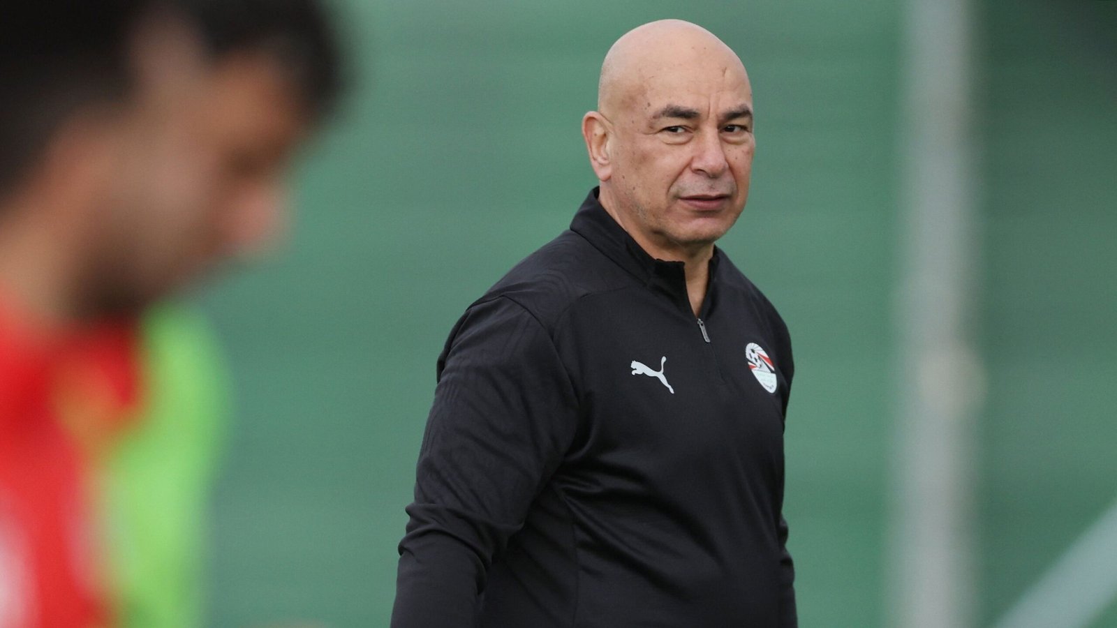 Egyptian football coach in a black Puma tracksuit during a training session, with a blurred teammate in red and a green background.