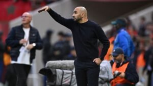 Walid Regragui gestures from the sidelines during a match, wearing a black top. The Moroccan flag is visible in the background.