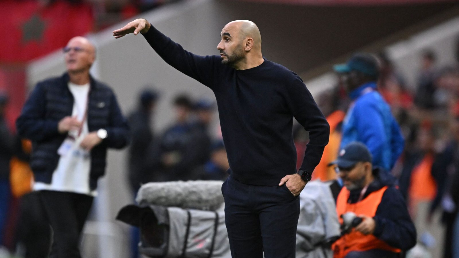 Walid Regragui gestures from the sidelines during a match, wearing a black top. The Moroccan flag is visible in the background.
