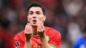 Un joueur marocain en maillot rouge de l'équipe nationale gesticule avec passion, reflétant l'intensité d'un match de la Coupe d'Afrique des Nations.