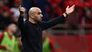 Walid Regragui gestures passionately from the sidelines during a match, wearing a black sweater, with a vibrant red crowd behind him.