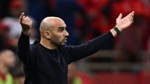 Walid Regragui gesturing passionately from the sidelines during a match, wearing a black sweater against a blurred red crowd backdrop.