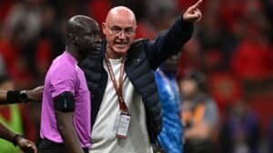 Referee in pink shirt and coach in a dark jacket argue passionately during a match, with fans in red in the background, highlighting tension.