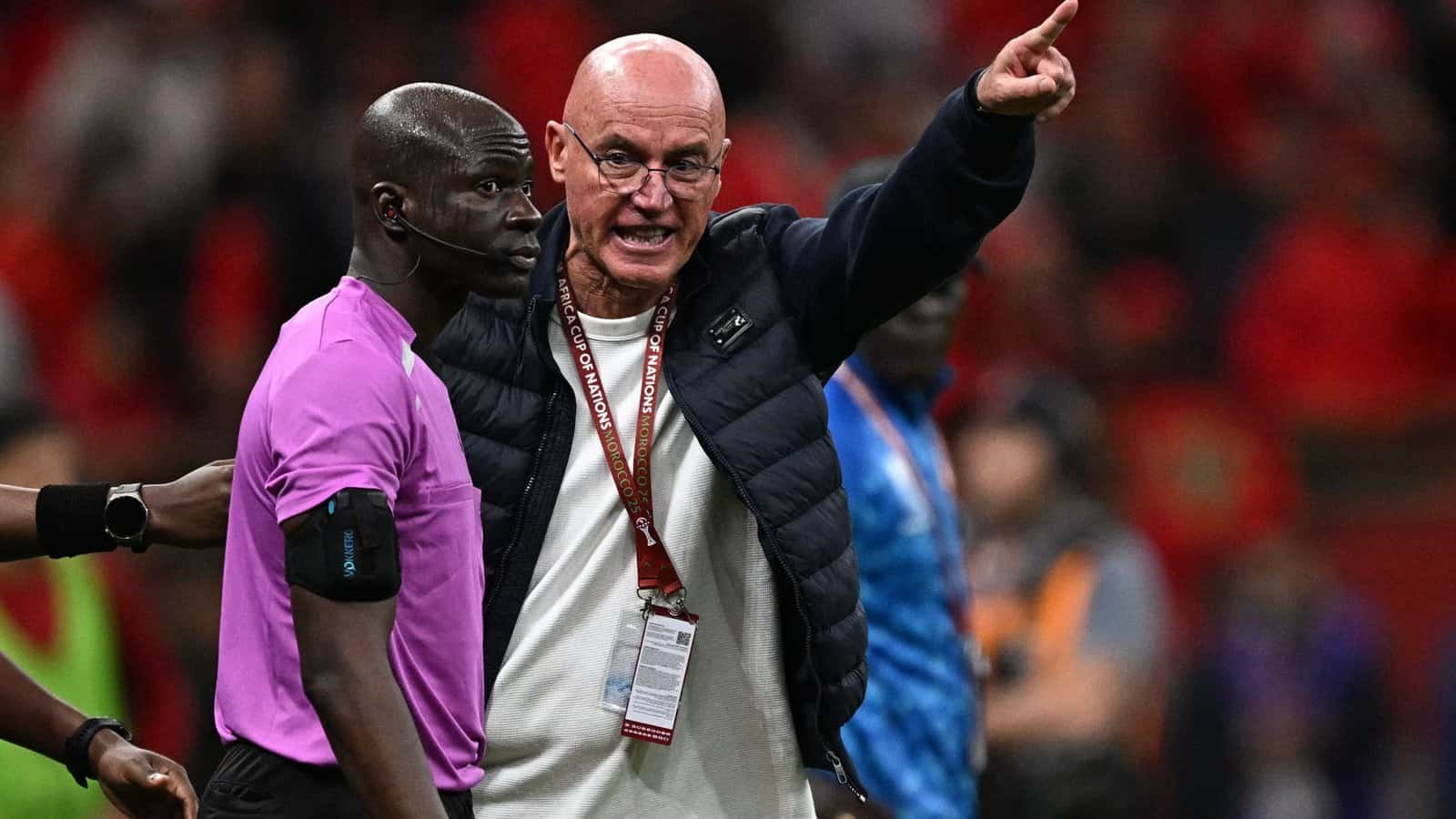 Referee in pink shirt and coach in a dark jacket argue passionately during a match, with fans in red in the background, highlighting tension.
