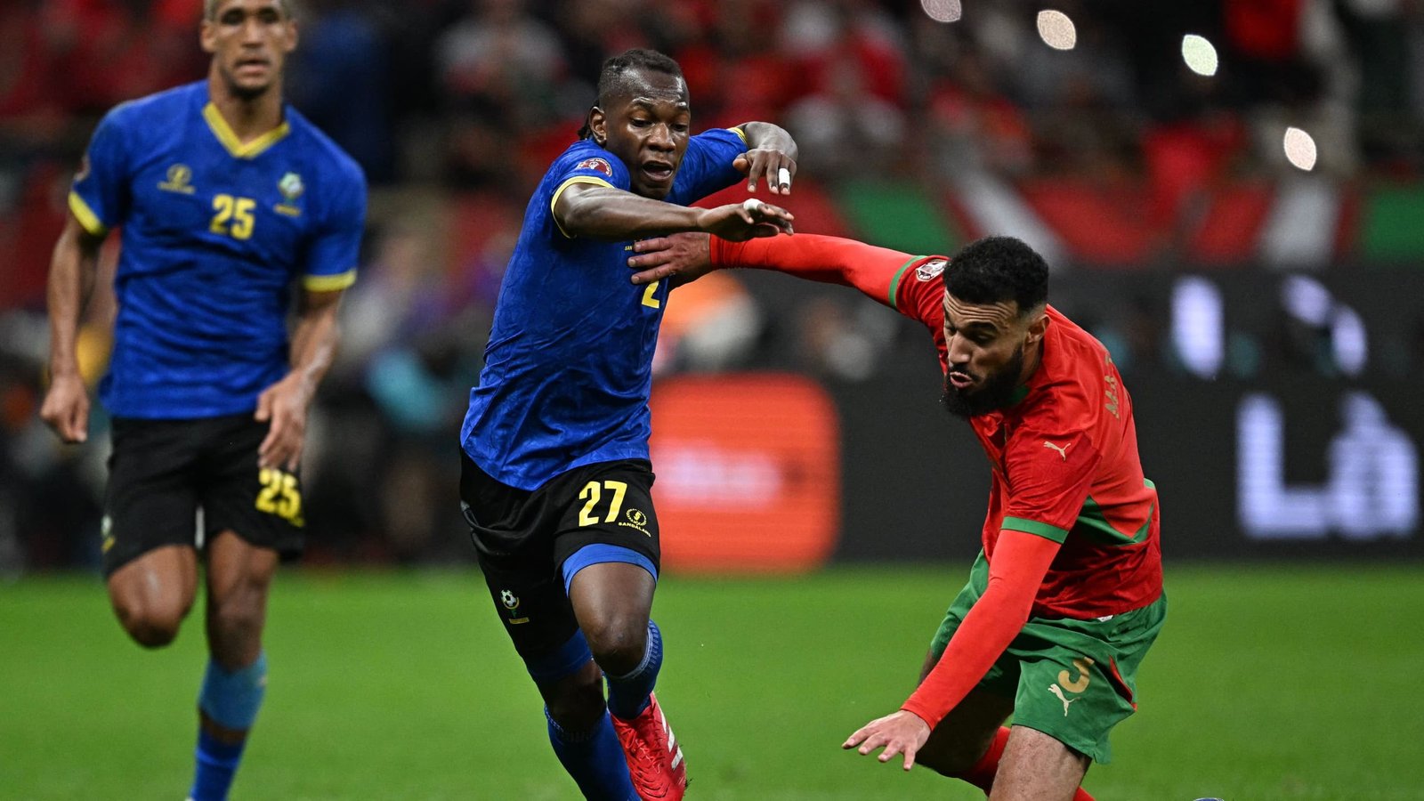Un joueur marocain conteste un adversaire portant un maillot rouge et vert éclatant lors d'un match intense contre la Tanzanie à la CAN.