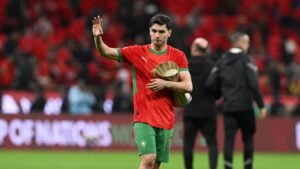 Achraf Hakimi walks off the pitch, holding a trophy, while wearing the Moroccan national team's red and green kit in a vibrant stadium.