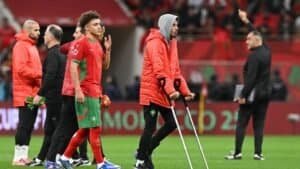 Les joueurs marocains en tenue rouge quittent le terrain, l'un d'eux s'appuyant sur des béquilles, tandis que le personnel de l'équipe et un entraîneur font des gestes en arrière-plan pendant un match.