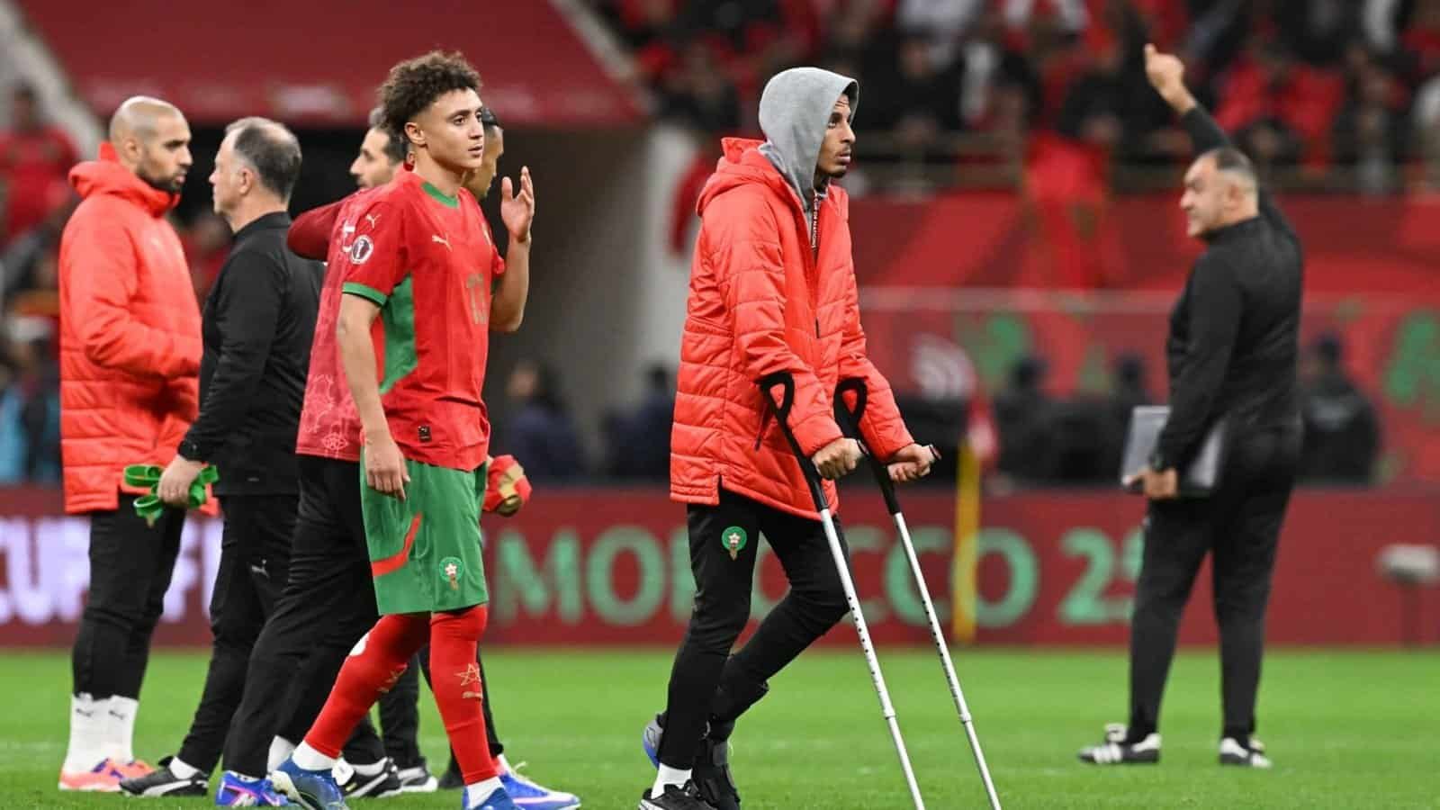 Les joueurs marocains en tenue rouge quittent le terrain, l'un d'eux s'appuyant sur des béquilles, tandis que le personnel de l'équipe et un entraîneur font des gestes en arrière-plan pendant un match.