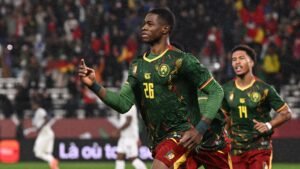 Le joueur numéro 26 du Cameroun célèbre un but dans un maillot vert vif agrémenté de touches jaunes et rouges, tandis que ses coéquipiers l'acclament en arrière-plan.