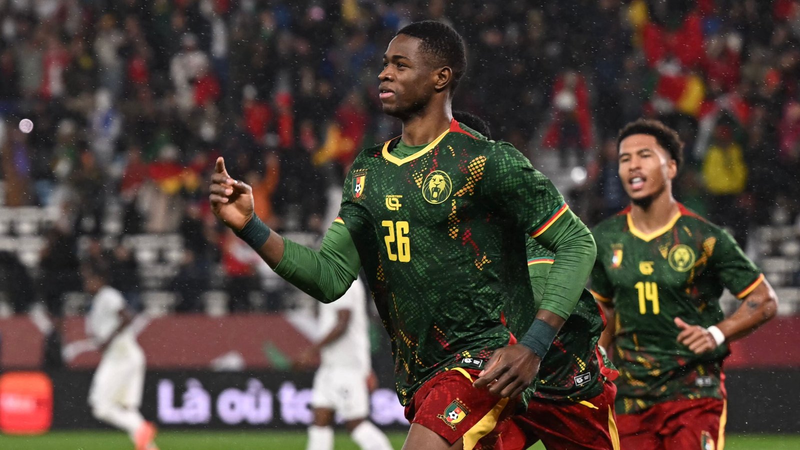 Le joueur numéro 26 du Cameroun célèbre un but dans un maillot vert vif agrémenté de touches jaunes et rouges, tandis que ses coéquipiers l'acclament en arrière-plan.
