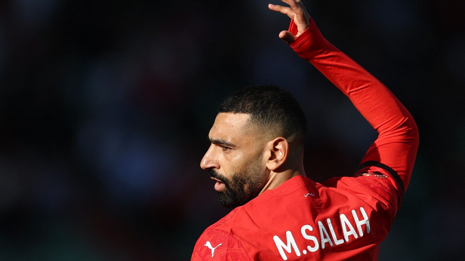 M. Salah gebaart zelfverzekerd terwijl hij een levendig rood teamtenue draagt, en toont zijn vastberadenheid tijdens een wedstrijd.