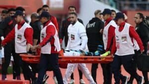 Le staff médical évacue un joueur blessé sur un brancard lors d'un match de football marocain, tandis que des coéquipiers en tenue rouge et blanche se tiennent à proximité.