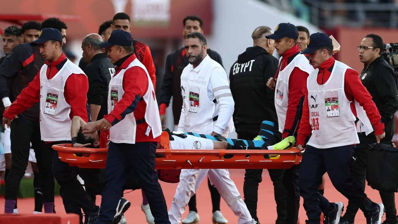 Le staff médical évacue un joueur blessé sur un brancard lors d'un match de football marocain, tandis que des coéquipiers en tenue rouge et blanche se tiennent à proximité.