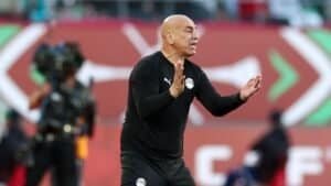 Entraîneur gesticulant avec passion en bord de terrain pendant un match de football, portant un survêtement noir avec le drapeau égyptien, sur fond animé.