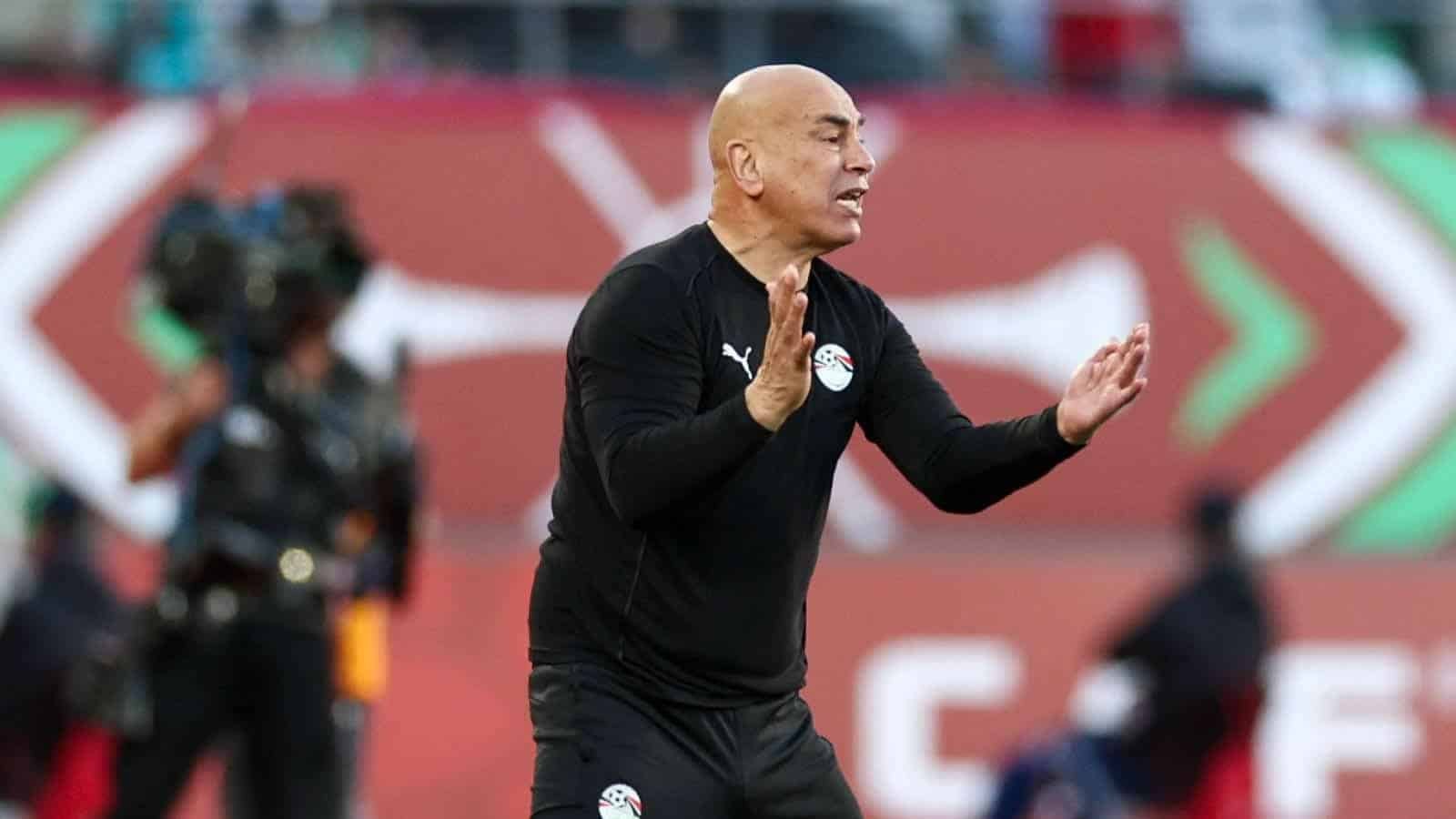 Entraîneur gesticulant avec passion en bord de terrain pendant un match de football, portant un survêtement noir avec le drapeau égyptien, sur fond animé.