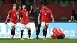 Les joueurs égyptiens célèbrent un but, portant des maillots rouges. Un joueur est à genoux sur le sol au premier plan, tandis qu'un arbitre observe le moment.