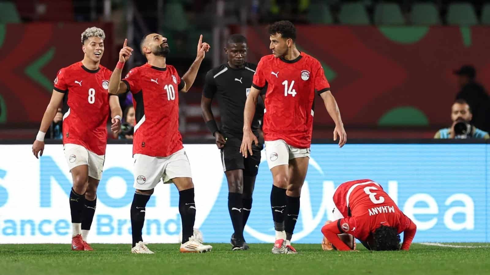 Les joueurs égyptiens célèbrent un but, portant des maillots rouges. Un joueur est à genoux sur le sol au premier plan, tandis qu'un arbitre observe le moment.