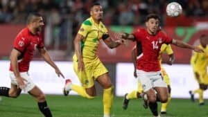 Des joueurs marocains et égyptiens rivalisent pour le ballon dans une scène de match dynamique, arborant des maillots rouge, jaune et vert, avec des supporters en arrière-plan.