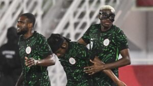 Nigerian players celebrate a goal, wearing their distinctive green jerseys with a patterned design, set against a stadium backdrop.