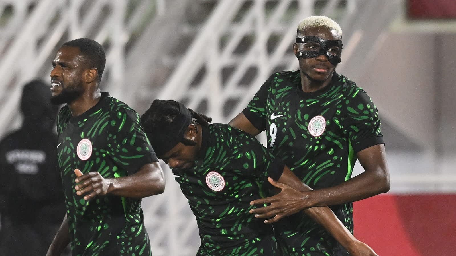 Nigerian players celebrate a goal, wearing their distinctive green jerseys with a patterned design, set against a stadium backdrop.