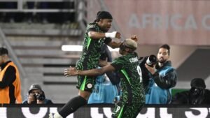 Nigeriaanse spelers die een doelpunt vieren tijdens de Afrika Cup, gekleed in levendige groene tenues met zwarte patronen, omringd door fotografen.