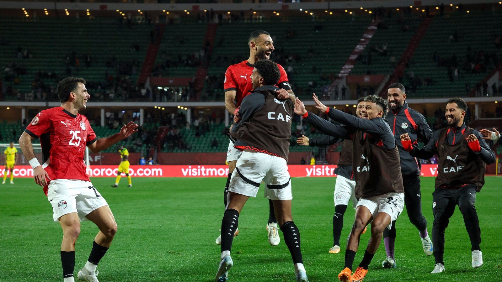 Egyptische spelers die gepassioneerd feestvieren op het veld, gekleed in rode en zwarte tenues, na een succesvolle wedstrijd, tegen een levendige stadionachtergrond.