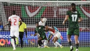 Marokkaanse en Nigeriaanse spelers die tijdens een gespannen moment in de wedstrijd om de bal strijden, met het doelnet en een levendige achtergrond zichtbaar.