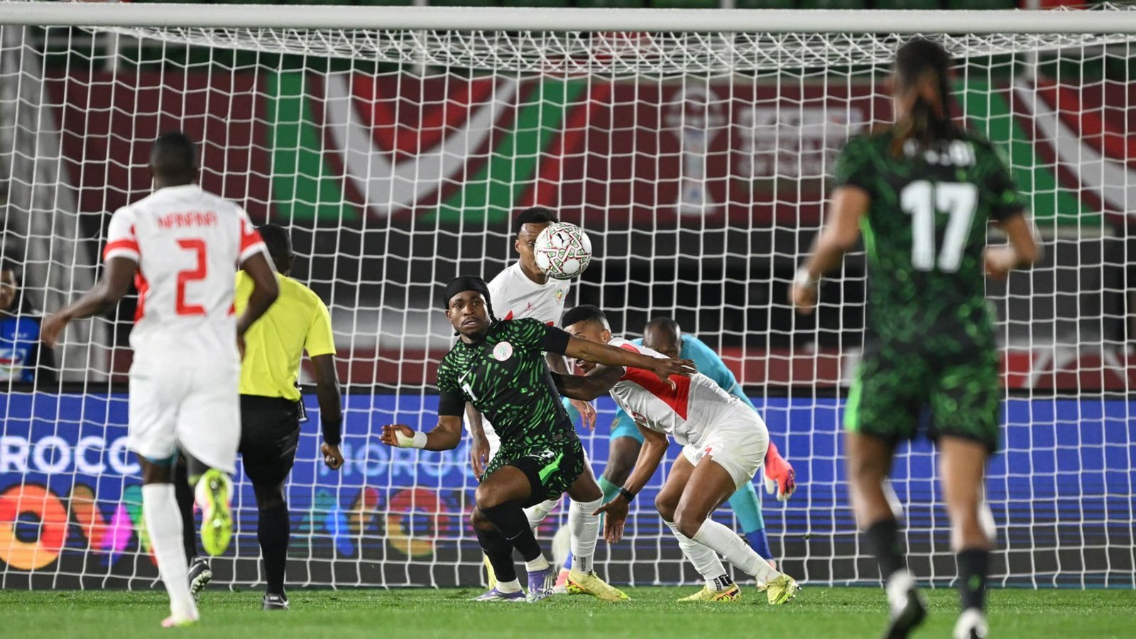 Marokkaanse en Nigeriaanse spelers die tijdens een gespannen moment in de wedstrijd om de bal strijden, met het doelnet en een levendige achtergrond zichtbaar.