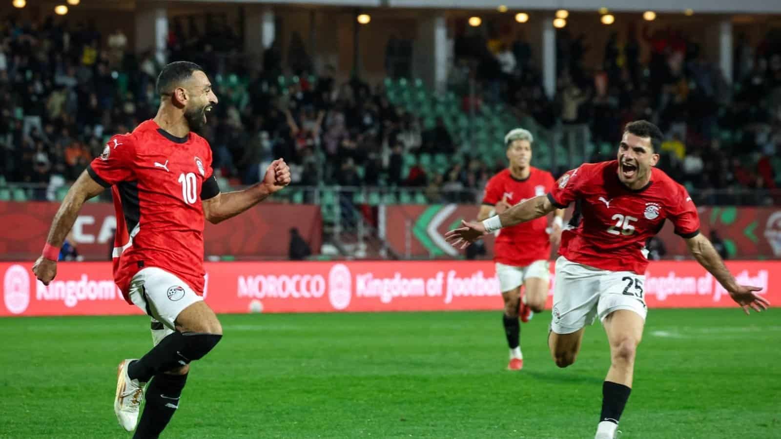 Des joueurs égyptiens célèbrent un but pendant un match, portant des maillots rouges avec les numéros 10 et 25, au milieu d'une foule vibrante.