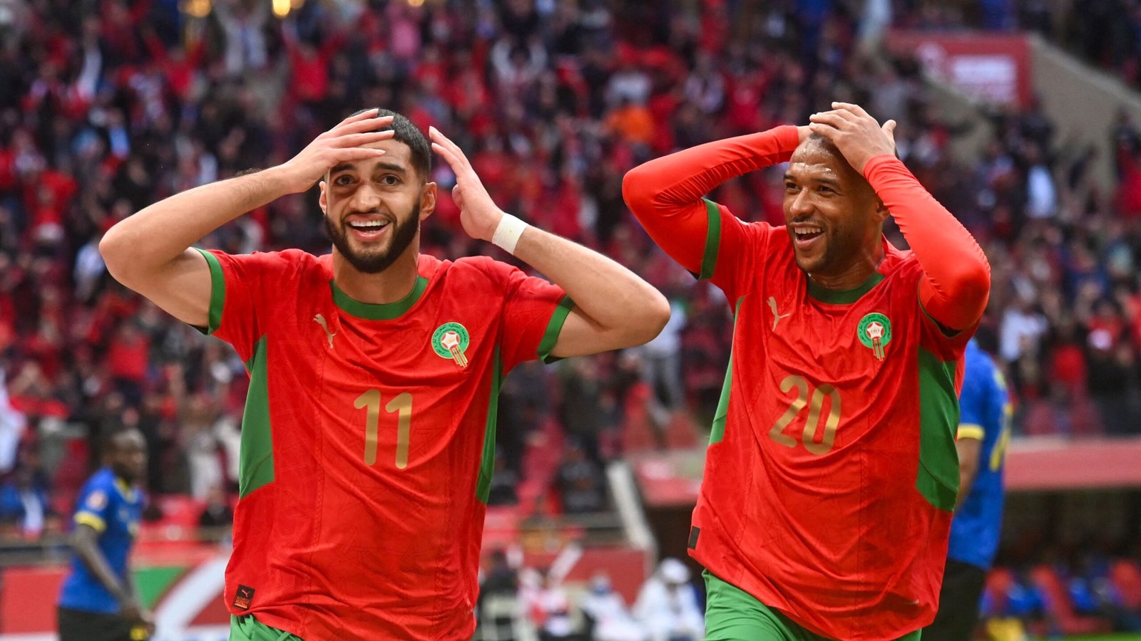 Joueurs marocains célébrant un but, vêtus de maillots vert et rouge, avec des expressions jubilatoires, sur fond de foule vibrante.