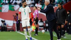Riyad Mahrez, portant le maillot blanc de l'Algérie avec des accents verts, écoute attentivement l'entraîneur Didier Deschamps lors d'un moment tendu sur la touche.