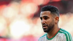 Riyad Mahrez dans le maillot de l'équipe marocaine, concentré avec une expression sérieuse, contre un arrière-plan flou de teintes rouges et jaunes vibrantes.