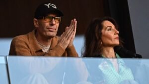 Deux spectateurs observent attentivement depuis une loge VIP du stade, un homme portant une casquette noire Y-3 et des lunettes, et une femme vêtue d'un chemisier vert à motifs.