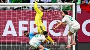 Le gardien en jaune effectue une parade, tandis que les joueurs algériens en blanc et bleu réagissent près du but lors d'un match intense de la Coupe d'Afrique.