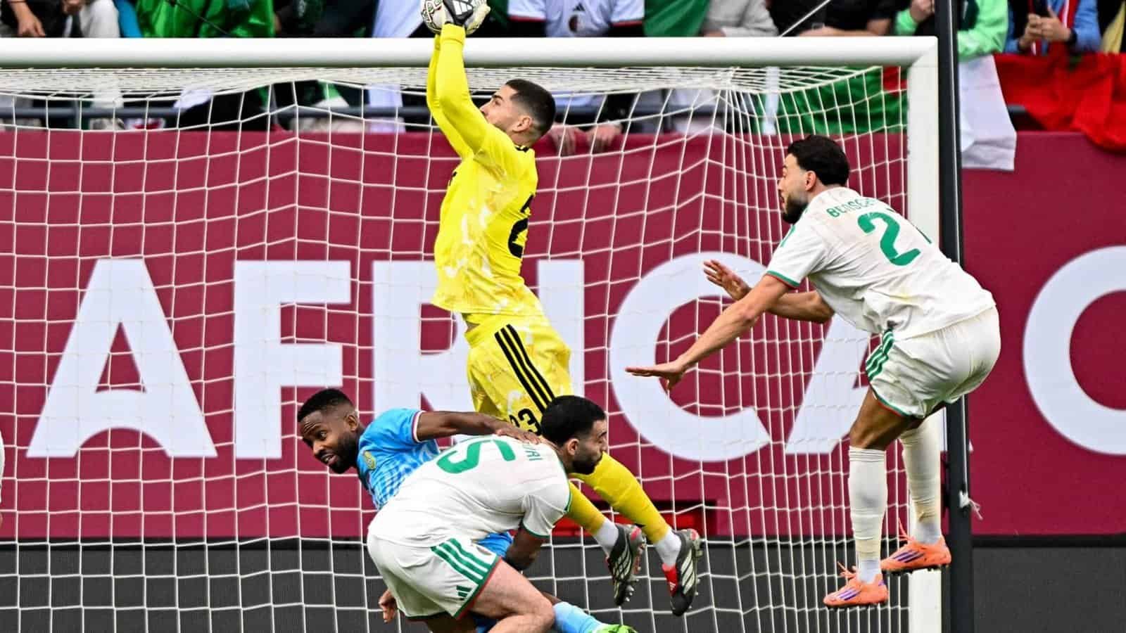 Le gardien en jaune effectue une parade, tandis que les joueurs algériens en blanc et bleu réagissent près du but lors d'un match intense de la Coupe d'Afrique.