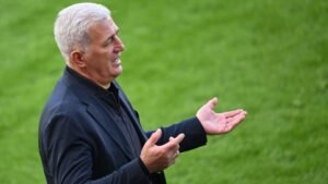 L'entraîneur gesticulant avec passion en bord de terrain, vêtu de noir sur un terrain d'un vert éclatant, transmettant une intense détermination pendant un match.