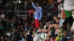 A fan stands proudly in a blue suit and red trousers, cheering among a crowd of supporters in a vibrant stadium atmosphere.