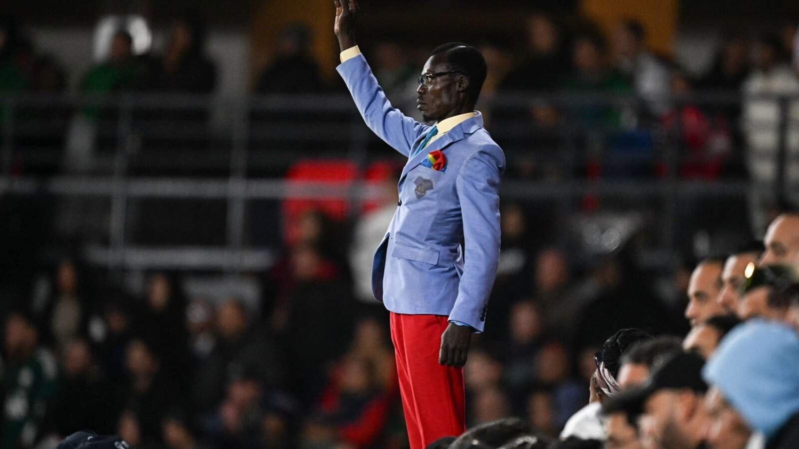A passionately dressed fan in a light blue suit and red trousers stands out amongst the crowd, waving enthusiastically during the match.