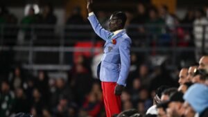 A stylish fan in a blue blazer and red trousers passionately cheers, raising his arm amid a lively crowd at a football match.